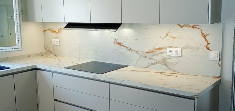 Encimera y frente de continuidad en porcelánico blanco con veta dorada, con placa de inducción negra y campana integrada bajo los muebles altos.